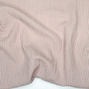 May include: A close-up of a fabric with a pale pink background and vertical, thin, dark red stripes. The fabric is slightly creased, creating soft shadows and texture. The stripes are evenly spaced and run the length of the fabric.
