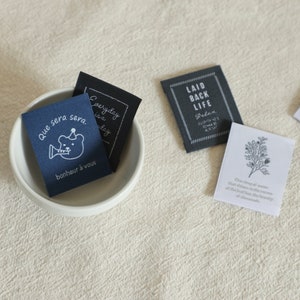 May include: Four fabric patches with different designs and text. The first patch is blue with a white bear playing a trumpet and the text "Que sera sera. bonheur à vous". The second patch is black with white text that says "Everyday is a day". The third patch is black with white text that says "Laid Back Life Relax. Everything's gonna be alright". The fourth patch is white with a grey floral design and the text "One sprig of "weed" that shines in the darkness of the leaf has the beauty of diamonds."