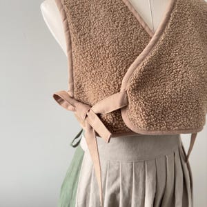 May include: A cropped, tan-colored vest made of a soft, textured material. The vest has a wrap-around design with a tie closure at the front. The edges are trimmed with a lighter tan fabric. The vest is displayed on a mannequin, paired with a pleated skirt.
