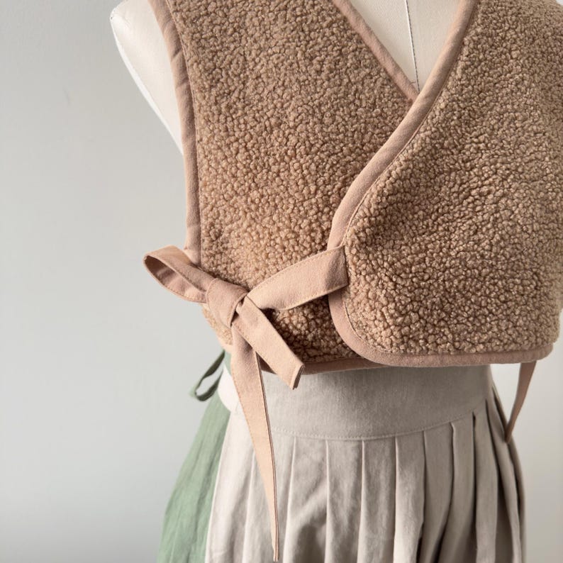 May include: A cropped, tan-colored vest made of a soft, textured material. The vest has a wrap-around design with a tie closure at the front. The edges are trimmed with a lighter tan fabric. The vest is displayed on a mannequin, paired with a pleated skirt.