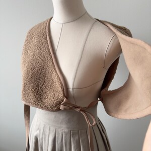 May include: A tan-colored, open-front shawl made of a textured, fuzzy material. The shawl has a smooth, light brown trim and ties at the front with a bow. The garment is displayed on a mannequin, showcasing its design and drape.