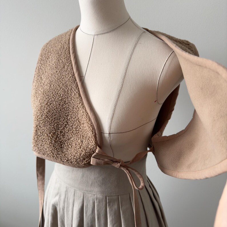 May include: A tan-colored, open-front shawl made of a textured, fuzzy material. The shawl has a smooth, light brown trim and ties at the front with a bow. The garment is displayed on a mannequin, showcasing its design and drape.