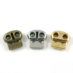 May include: Three toggle cord locks in different finishes: antique brass, silver, and gold. Each lock has two holes and a rectangular base.
