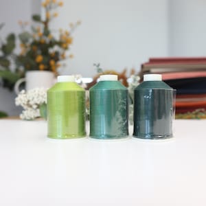 May include: Three spools of thread in shades of green. The spools are clear plastic and the thread is wound tightly. The thread colours are a light green, a medium green, and a dark green.
