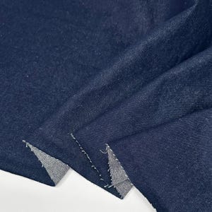May include: A length of dark navy blue denim fabric with a visible twill weave texture. The material is folded to reveal the lighter blue underside at the raw, unfinished edges, showcasing the classic construction of this durable textile.