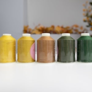 May include: Five spools of thread in various shades of yellow, brown, and green. The spools are arranged in a row from left to right.