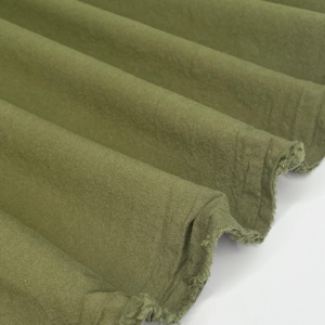 May include: Close-up of olive green fabric with a textured appearance. The fabric is folded in diagonal lines, creating shadows and highlighting the texture. The edges of the fabric are frayed.