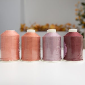 May include: Four spools of thread in shades of pink, orange, and red. The spools are clear plastic and the thread is wound tightly.