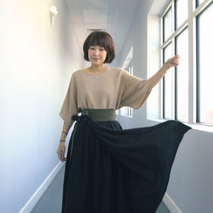 Pode incluir: Uma mulher vestindo uma blusa bege com uma faixa verde e uma saia preta de enrolamento. Ela est&aacute; em p&eacute; em um corredor com uma janela no fundo.