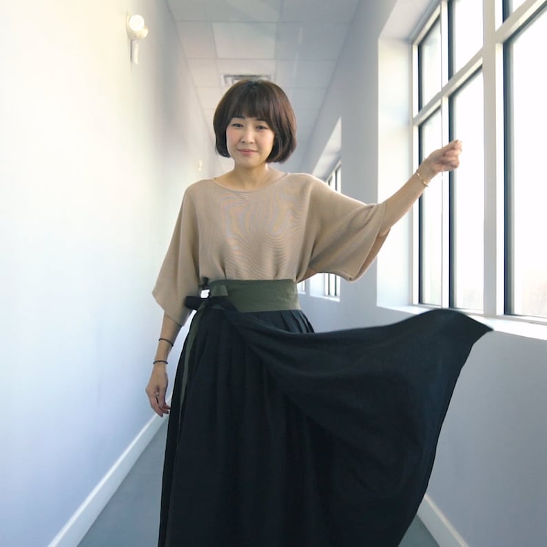 Pode incluir: Uma mulher vestindo uma blusa bege com uma faixa verde e uma saia preta de enrolamento. Ela est&aacute; em p&eacute; em um corredor com uma janela no fundo.