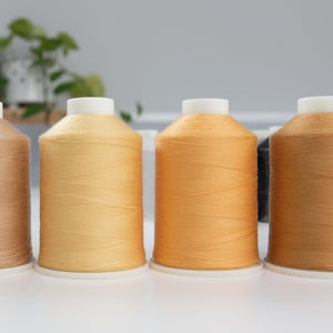 Peut inclure: Une rangée de quatre bobines cylindriques de fil à coudre dans des tons variés de beige, de brun clair et d'orange. Chaque bobine présente un sommet et une base en plastique blanc, avec le fil enroulé serré autour du centre de manière uniforme.