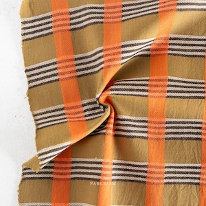 May include: A folded textile with a plaid pattern. The fabric features alternating stripes of orange, brown, and white. The background colour is a muted gold. The fabric is slightly wrinkled, showing the texture of the material.