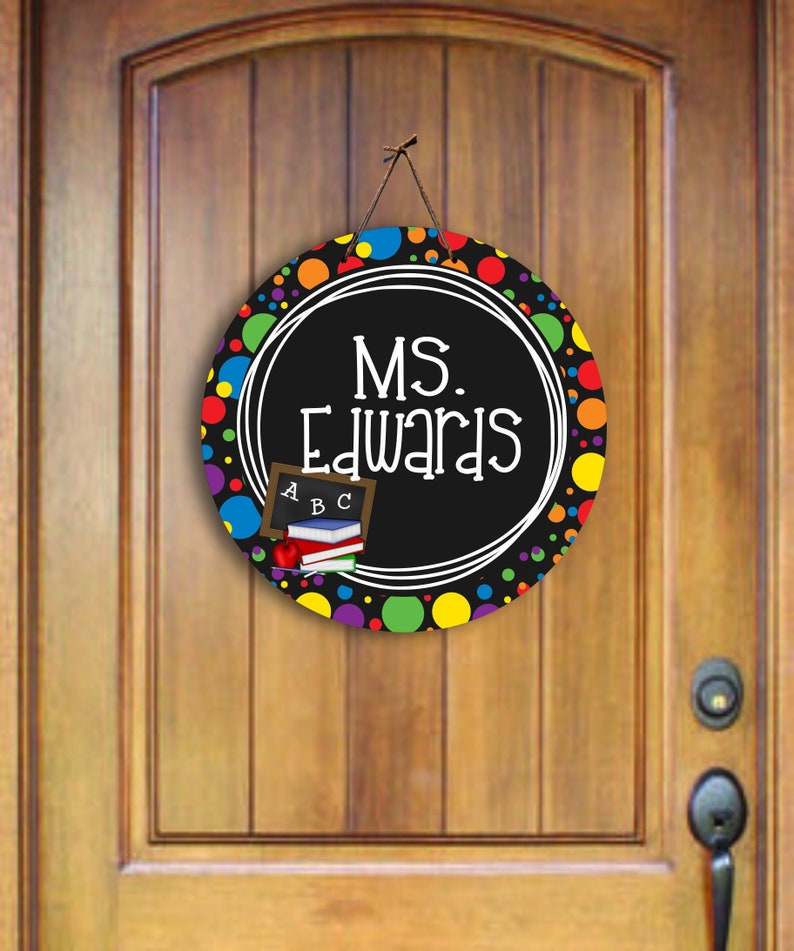 May include: A black and white round door hanger with colourful polka dots. The text "MS. Edwards" is written in a white circle on the door hanger. There is a small chalkboard with the letters "ABC" and a stack of books on the door hanger.