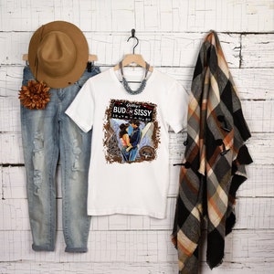Puede incluir: Una camiseta blanca con un gráfico que dice "Gilley's Bud & Sissy 19-Texas-80" con una pareja. La camiseta se muestra con vaqueros azules desgastados, un sombrero marrón, un adorno floral, un collar y una bufanda a cuadros.
