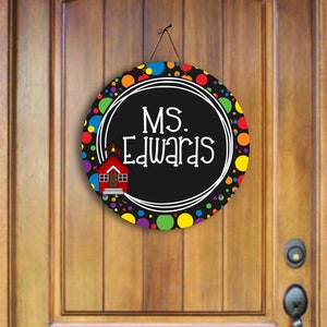 May include: A round door hanger with a black and white background and a colourful polka dot border. The centre features a white circle with the text "MS. Edwards" and a small red schoolhouse illustration.