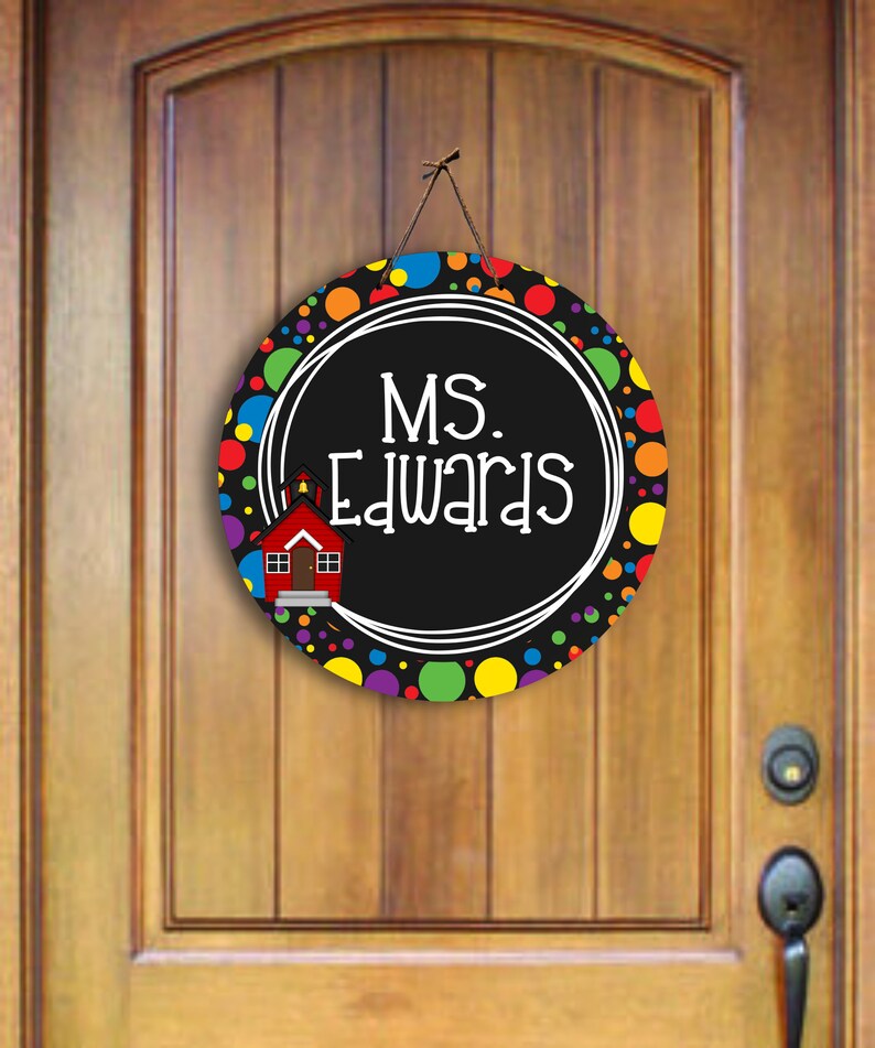 May include: A round door hanger with a black and white background and a colourful polka dot border. The centre features a white circle with the text "MS. Edwards" and a small red schoolhouse illustration.
