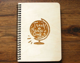May include: A wooden spiral-bound notebook with a light wood cover. The cover features a globe design with the phrase "Travel is always a Good idea" etched in a brown color. The notebook is on a wooden surface.