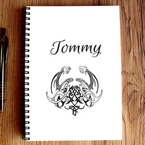May include: A white spiral bound notebook with the name "Tommy" written in black cursive on the front cover. The notebook has a black and white illustration of two dragons with their tails intertwined.