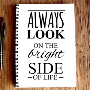 May include: A white notebook with a black spiral binding. The notebook has the text "ALWAYS LOOK ON THE bright SIDE -OF LIFE- " written on it.