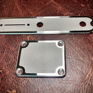 Può includere: Due piastre per chitarra in metallo argentato. La piastra più grande è rettangolare con bordi arrotondati e due fori. La piastra più piccola è quadrata con angoli arrotondati e quattro fori.