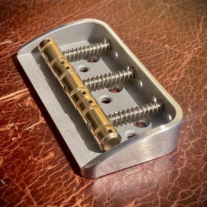 May include: A silver guitar bridge with three springs and a gold barrel. The bridge is rectangular with rounded edges. The barrel has several holes. It sits on a brown, textured surface.