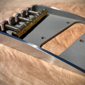 May include: A silver guitar bridge with six individual saddles and a brass bridge bar. The bridge is designed for a six-string guitar.