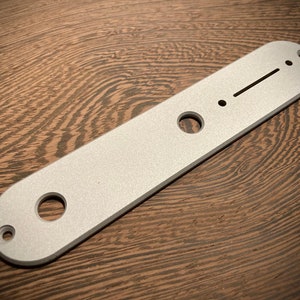 Puede incluir: Una pieza metálica plateada para guitarra, de forma rectangular con extremos redondeados. Tiene tres agujeros de diferentes tamaños y una serie de marcas grabadas. La pieza está sobre un fondo de vetas de madera oscura.