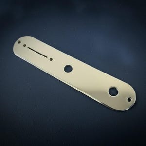 May include: A gold-colored metal guitar control plate with a rectangular shape and rounded ends. It features a long, thin slot, two small holes, and two larger circular holes. The plate is reflective and sits on a dark surface.