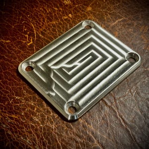 Könnte beinhalten: Eine quadratische, gebürstete Metallplatte mit einem eingelassenen Labyrinth-Design. Die Platte hat abgerundete Ecken und vier gleichmäßig verteilte Löcher. Sie liegt auf einer strukturierten, braunen Lederoberfläche.
