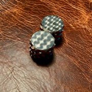 Puede incluir: Dos dados de metal plateados con una superficie texturizada y martillada. Los dados son redondos y tienen un borde ligeramente elevado.