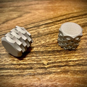 May include: Two small, silver-colored, geometric objects with a textured, layered design. The objects are cylindrical with a flat top and bottom, and are resting on a wooden surface.