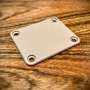 May include: A silver-coloured metal plate with four holes for screws. The plate has a textured surface.