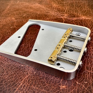 Pode incluir: Uma ponte de guitarra prateada com pinos de afinação e molas douradas. A ponte é projetada para uma guitarra de seis cordas.