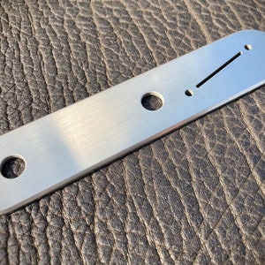 Könnte beinhalten: Eine Gitarrenbrückenplatte aus gebürstetem Metall mit rechteckiger Form und abgerundeten Enden. Sie hat einen langen, abgewinkelten Schlitz und mehrere kreisförmige Löcher zur Montage und Saitenausrichtung. Die Platte wird vor einem strukturierten, dunklen Hintergrund gezeigt.