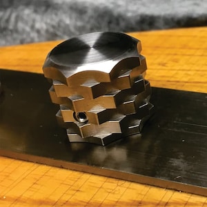 Puede incluir: Un pomo de metal plateado con forma hexagonal y un acabado liso y pulido. El pomo tiene un pequeño agujero en el centro.