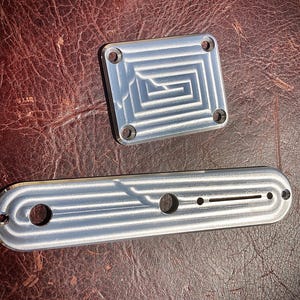 May include: Two silver metal guitar parts, a rectangular plate with a maze-like design and a long, curved plate with holes. The parts are on a textured, brown surface. The parts have a machined finish.