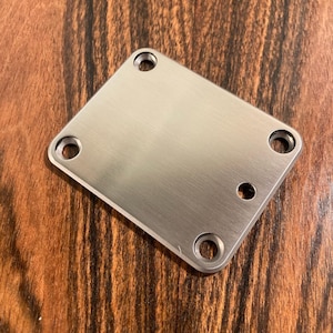 Op de afbeelding: Een zilveren metalen gitaarplaat met vier schroefgaten en een afgeronde rand.