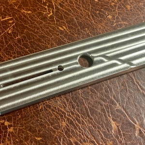 May include: A silver-coloured metal guitar component with a series of parallel grooves and several holes. The rectangular piece has rounded ends and a slot. The metal is set against a brown leather background.