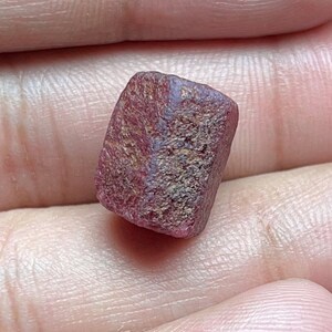 May include: A small, rough, rectangular-shaped gemstone with a deep reddish-pink hue and a textured, matte surface. The stone displays natural crystalline facets and variations in color intensity across its surface, appearing opaque and unpolished.