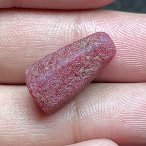 May include: A small, raw, unpolished ruby gemstone with a deep red color and a rough, textured surface. The stone has an elongated, slightly tapered shape with natural facets and irregular edges, showcasing its authentic, untreated mineral form.