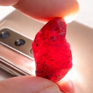 Könnte beinhalten: Ein roher, ungeschliffener Rubin-Edelstein, der zwischen Fingern gehalten wird. Der Rubin hat eine tiefe, leuchtende rote Farbe mit einer strukturierten Oberfläche. Der Hintergrund zeigt ein goldfarbenes Smartphone.