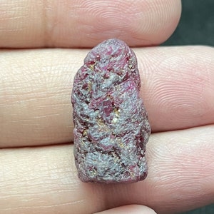 May include: A rough-cut ruby gemstone, displaying a deep red hue with a textured surface. The natural gemstone has a slightly elongated shape and is held between two fingers.