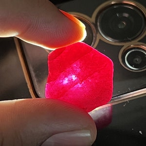 May include: A vibrant, translucent red gemstone with an irregular, somewhat hexagonal shape. The stone exhibits a deep crimson hue and is illuminated from behind, revealing internal textures and light diffusion throughout its crystalline structure.