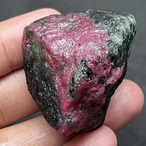 Könnte beinhalten: Ein rohes, unpoliertes Mineralexemplar mit einem markanten, leuchtend magentafarbenen kristallinen Bereich, der in eine dunkle, texturierte Matrix aus dunkelgrünem und schwarzem Gestein eingebettet ist, mit natürlichen Kanten und rauer Oberfläche.