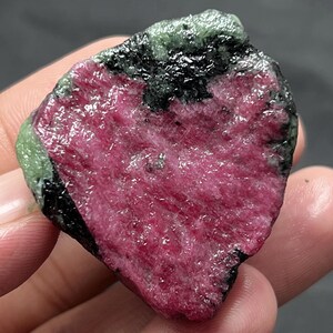 Könnte beinhalten: Ein rohes, unpoliertes Mineralexemplar mit einem leuchtend magentafarbenen Zentrum, das von Flecken aus tiefschwarzen und hellgrünen Mineralablagerungen umgeben ist. Der Stein hat eine unregelmäßige, abgerundete Form mit einer texturierten Oberfläche.