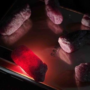 May include: Several rough, irregular, pinkish-red gemstone crystals are arranged on the reflective, metallic surface of a smartphone. One crystal is illuminated from beneath, emitting a bright, glowing red light, while the others remain dark.