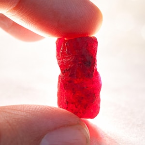 May include: A raw, uncut ruby gemstone, held between two fingers. The ruby is a deep, vibrant red color with a rough, textured surface. The gemstone is approximately 1 inch in length, showcasing its natural, unpolished form.