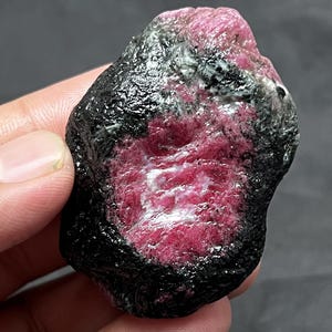 May include: A raw, unpolished mineral specimen featuring a deep magenta-pink core surrounded by a rough, textured black outer layer. The stone has an irregular, organic shape with a glossy, crystalline centre that contrasts with the dark exterior.