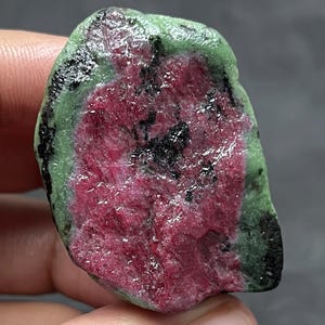 Peut inclure: Une tranche brute et polie de rubis zoïsite présentant un centre magenta-rose vibrant entouré d'une bordure verte épaisse et texturée, avec des inclusions noires dispersées sur toute la surface de la pierre, affichant une forme naturelle irrégulière.