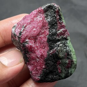 Op de afbeelding: Een ruw, ongepolijst mineraal exemplaar met een levendig magenta-roze middengedeelte dat contrasteert met diepzwarte en gedempte groene mineraalafzettingen. De steen heeft een onregelmatige, grillige vorm met een getextureerd, kristallijn oppervlak.